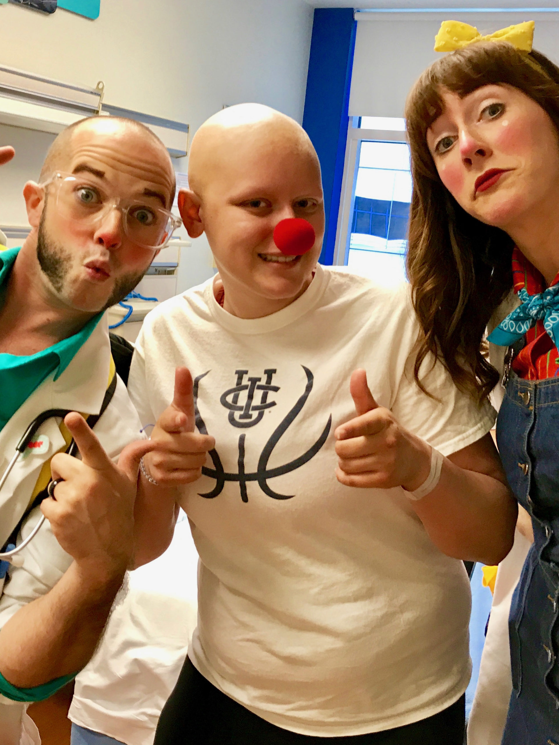 Sydney wearing a red nose posing with two clowns in her hospital room